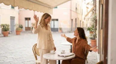 Dwie kobiety witają się serdecznie przy stoliku włoskiej kawiarni na słonecznej piazzy - jedna macha ręką, druga rozkłada ramiona, obok filiżanki espresso