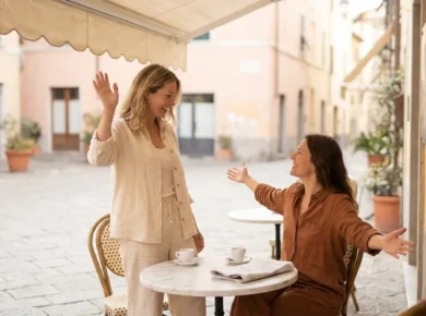 Dwie kobiety witają się serdecznie przy stoliku włoskiej kawiarni na słonecznej piazzy - jedna macha ręką, druga rozkłada ramiona, obok filiżanki espresso