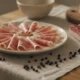 Plastry zwijanej pancetty arrotolata na jasnym ceramicznym talerzu, obok kawałki płaskiej pancetty tesy z warstwami mięsa i tłuszczu oraz ziarna czarnego pieprzu na lnianej ściereczce na drewnianym stole