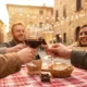 Przyjaciele wznoszą toast kieliszkami czerwonego wina i prosecco przy stole z kraciastym obrusem na kamiennym placu włoskiego miasteczka, w złotym świetle popołudnia